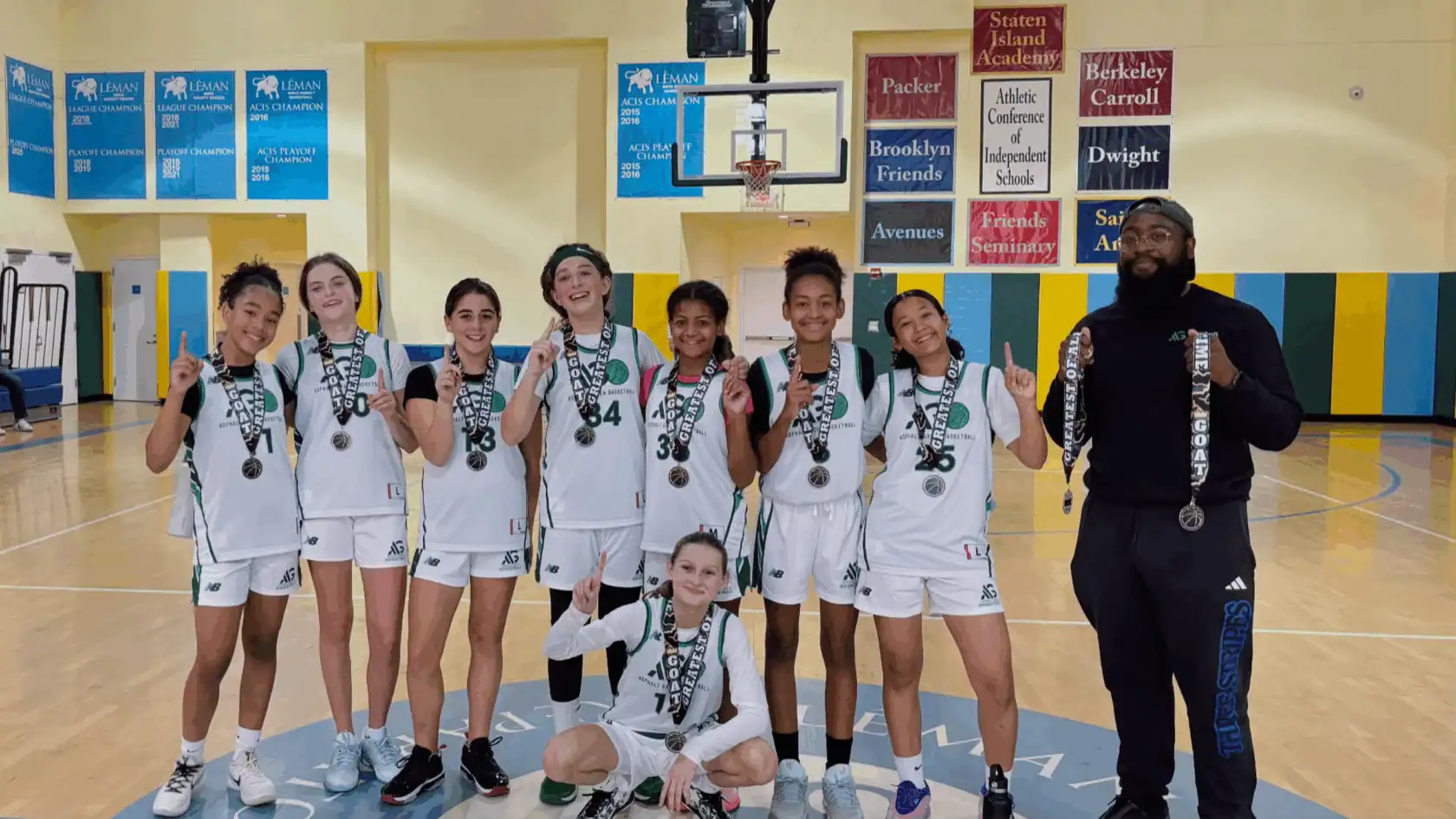 A girls’ basketball team in white uniforms stands in a gym, smiling and holding up medals, with their coach standing to the right.