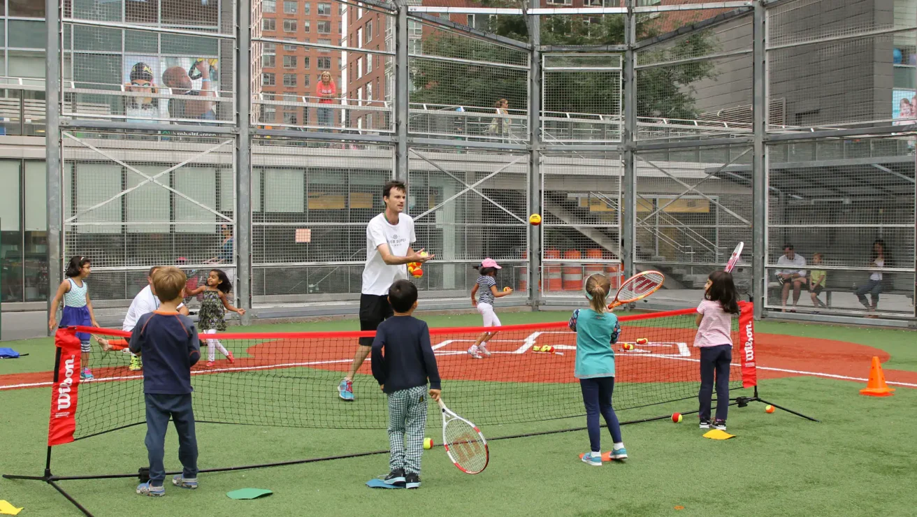 A coach tosses tennis balls to a group of young children holding rackets on an outdoor court. Urban buildings and spectators are visible in the background.