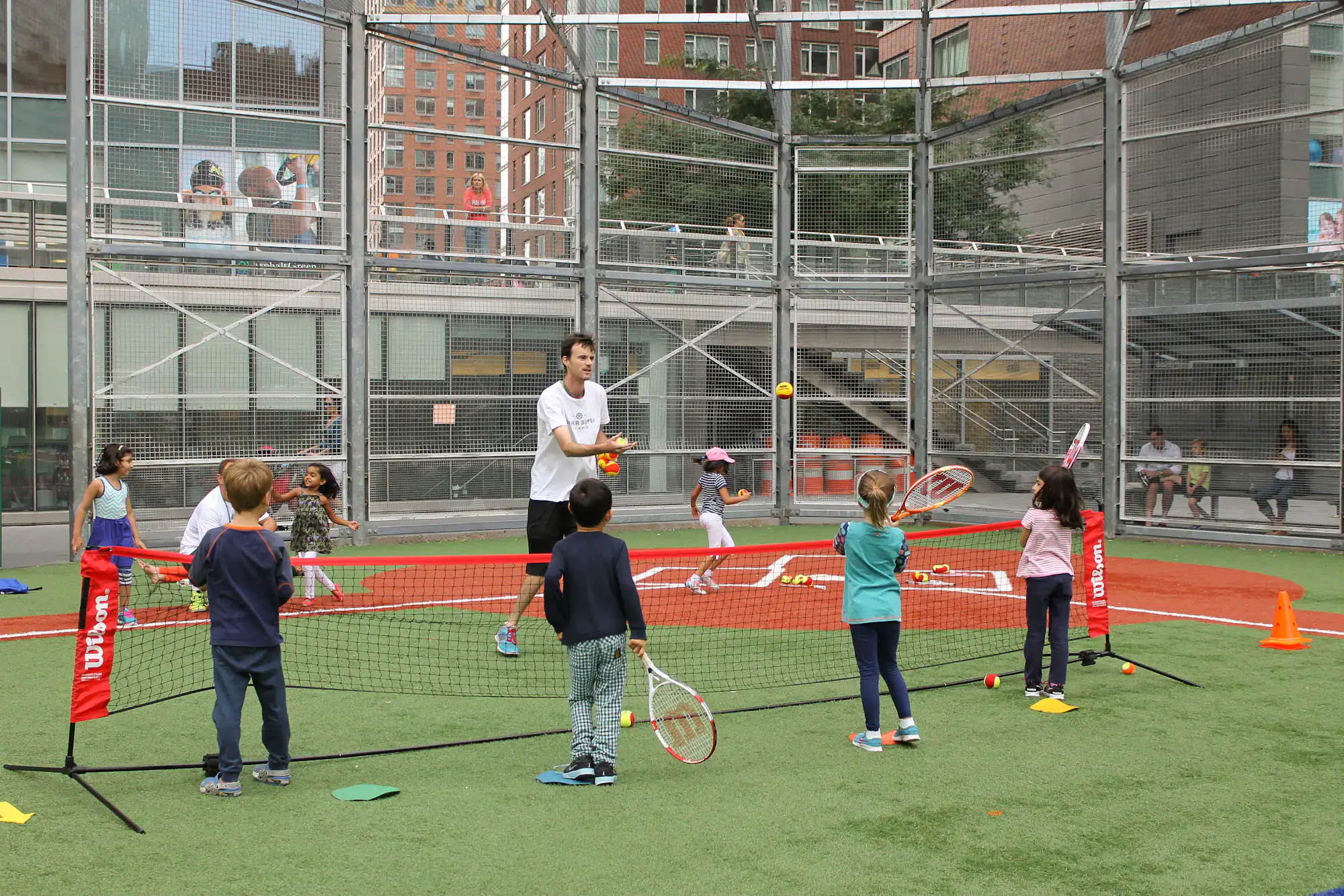 A coach tosses tennis balls to a group of young children holding rackets on an outdoor court. Urban buildings and spectators are visible in the background.