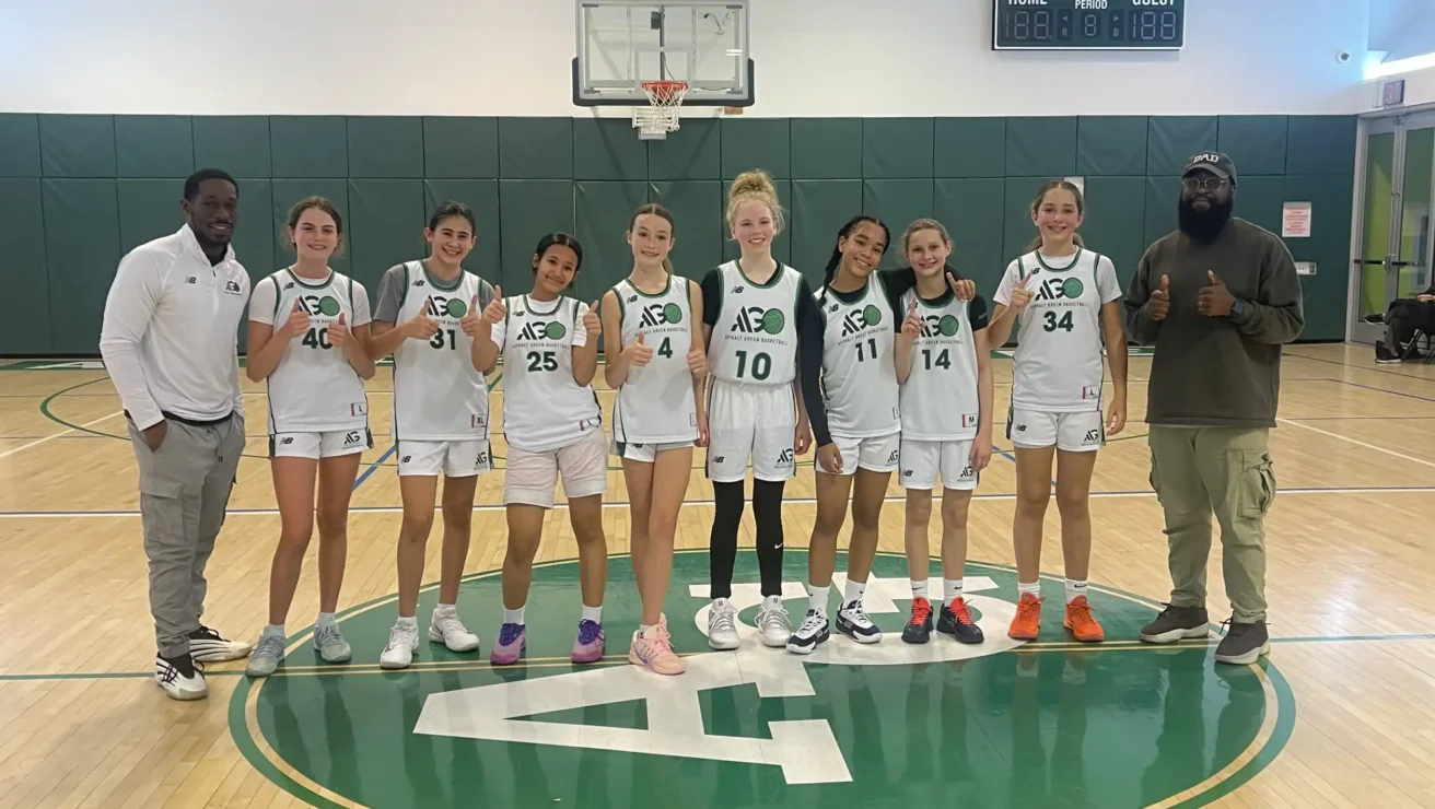 A girls’ basketball team in white and green uniforms poses on a gym court with two coaches, all holding up one finger in front of a basketball hoop.