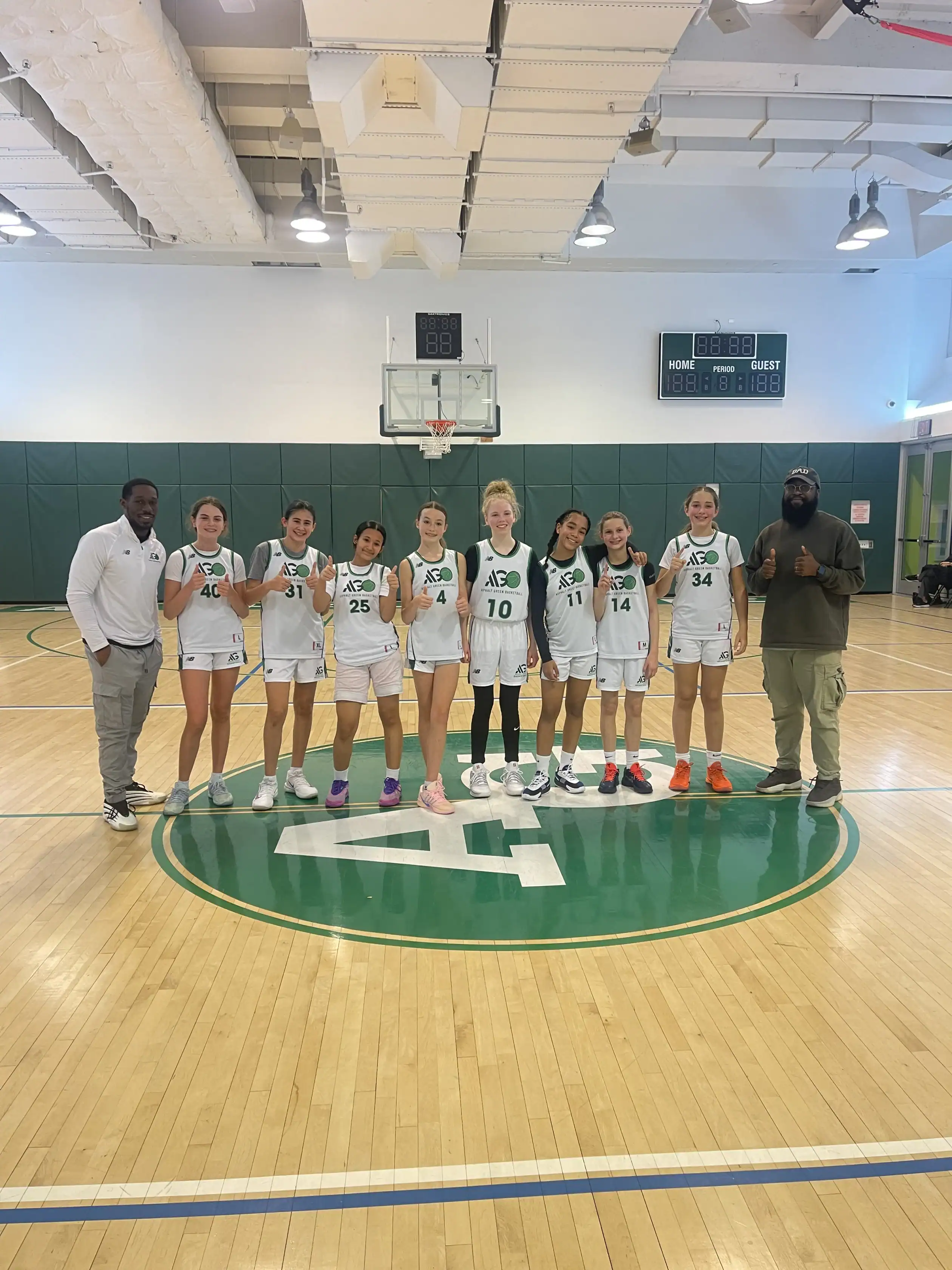 A girls’ basketball team in white and green uniforms poses on a gym court with two coaches, all holding up one finger in front of a basketball hoop.