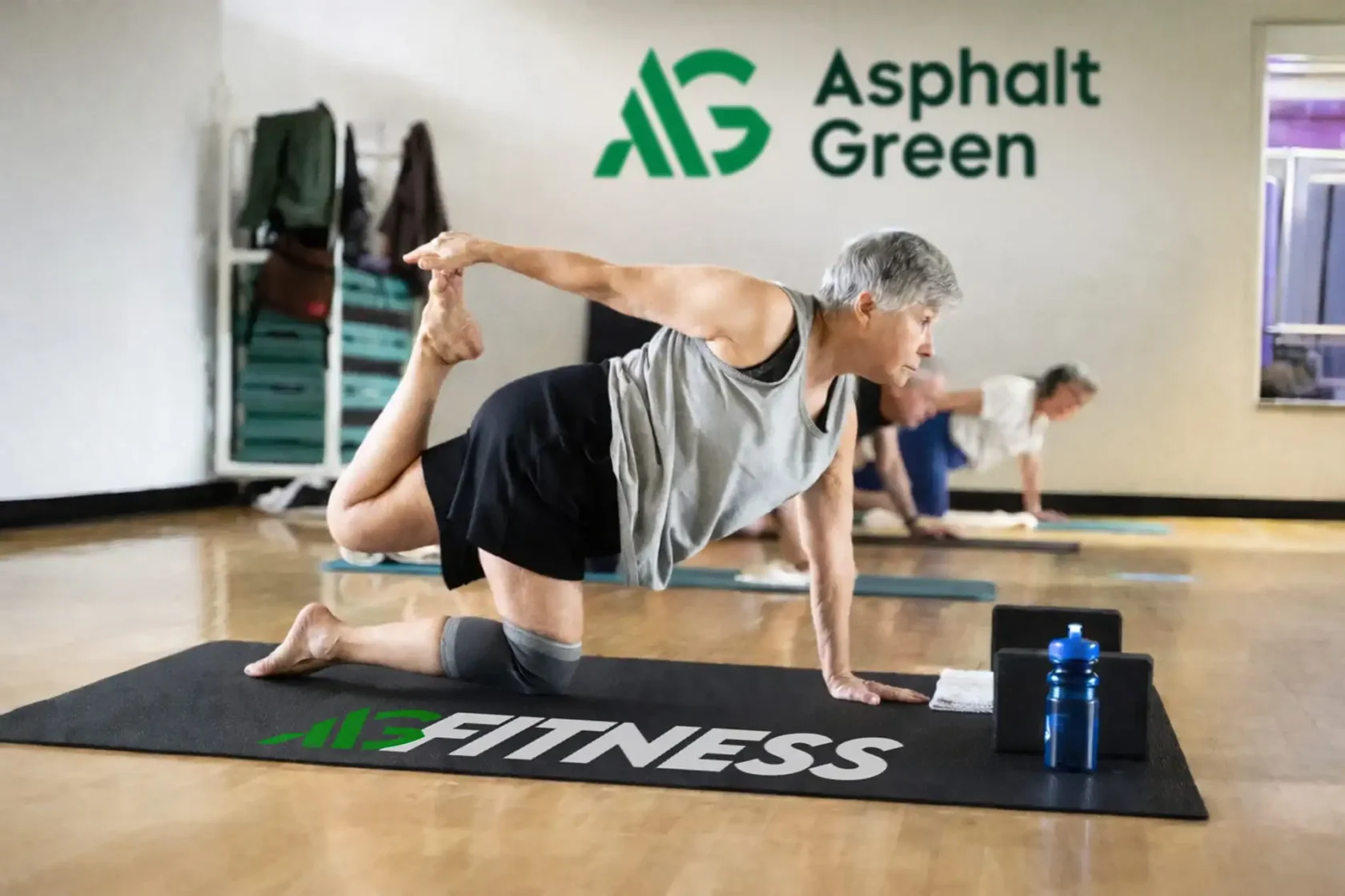 Older adults participate in a fitness class, performing stretching exercises on mats in a gym with "Asphalt Green" branding on the wall.