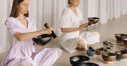 Two women in light dresses sit cross-legged on the floor, playing singing bowls with mallets, surrounded by various bowls, in a bright, minimalist room.