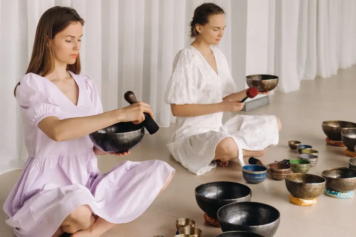 Two women in light dresses sit cross-legged on the floor, playing singing bowls with mallets, surrounded by various bowls, in a bright, minimalist room.