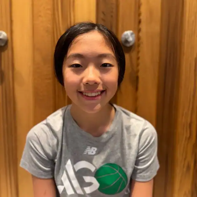 A young person with short black hair smiles at the camera, wearing a gray t-shirt with a green basketball graphic, seated against a wooden background.