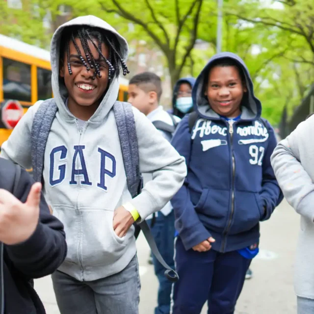 A group of school children wearing hoodies and backpacks stand outdoors near a school bus, some smiling and some wearing face masks.