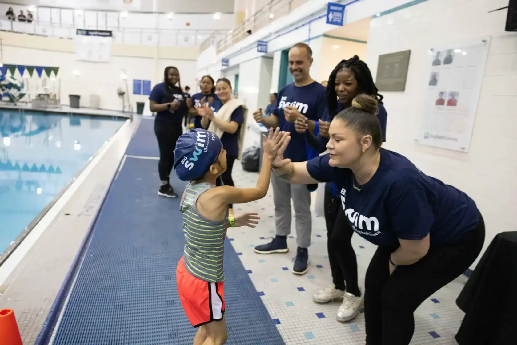 A child in swim gear high-fives an instructor by an indoor pool, while other adults and children watch and applaud in the background.