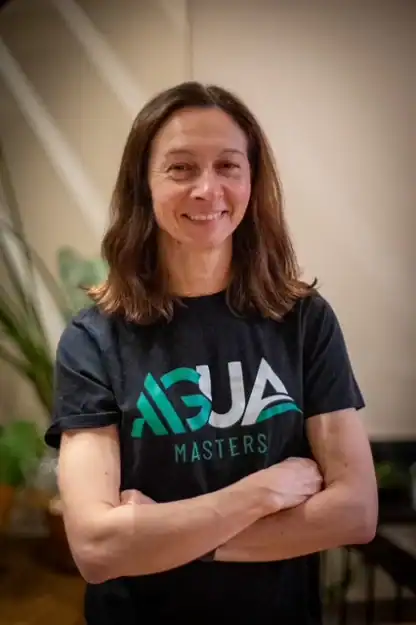 A person with shoulder-length brown hair stands indoors, smiling with arms crossed, wearing a black "AGUA Masters" t-shirt.