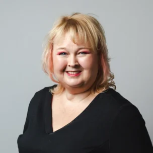 A woman with blonde hair and light makeup smiles at the camera, wearing a black V-neck top against a plain gray background.