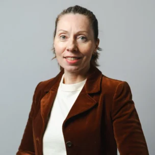 A woman with light skin and blue eyes, wearing a brown blazer over a white top, stands against a plain gray background, smiling at the camera.