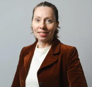 A woman with light skin and blue eyes, wearing a brown blazer over a white top, stands against a plain gray background, smiling at the camera.