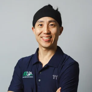 A person wearing a navy polo shirt and black headband stands with arms crossed, smiling at the camera against a plain background.