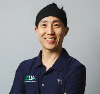 A person wearing a navy polo shirt and black headband stands with arms crossed, smiling at the camera against a plain background.