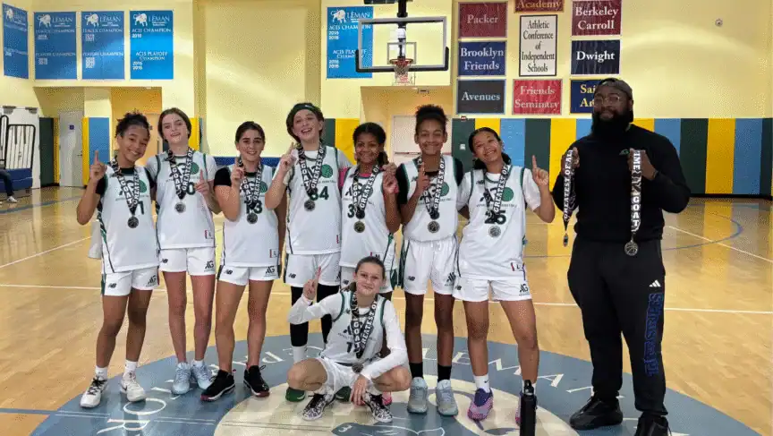 Eight basketball players and a coach pose on a gym court wearing medals, with trophies and school banners visible in the background.