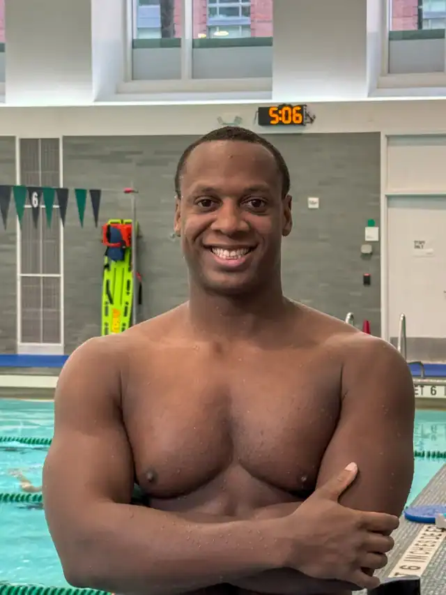 A man stands shirtless with crossed arms and smiles at an indoor swimming pool, with pool equipment and a clock in the background.