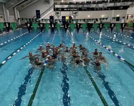 A group of people pose together in the middle of an indoor swimming pool, smiling, with triangular banners overhead spelling “GO AS PHALT GREEN,” celebrating National Girls and Women in Sports Day.