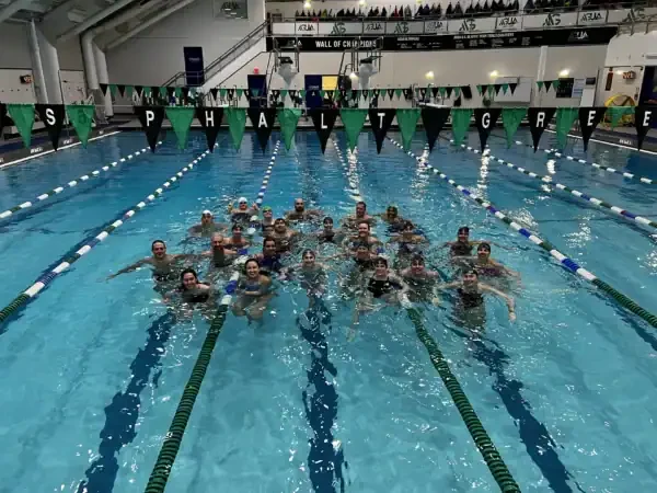 A group of people pose together in the middle of an indoor swimming pool, smiling, with triangular banners overhead spelling “GO AS PHALT GREEN,” celebrating National Girls and Women in Sports Day.