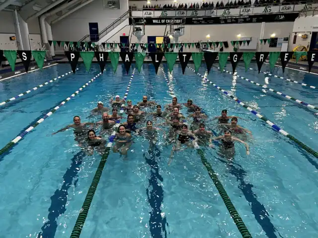 A group of swimmers posing together in an indoor swimming pool, with green and black pennant flags overhead and "ASPHALT GREEN" spelled out on the flags.