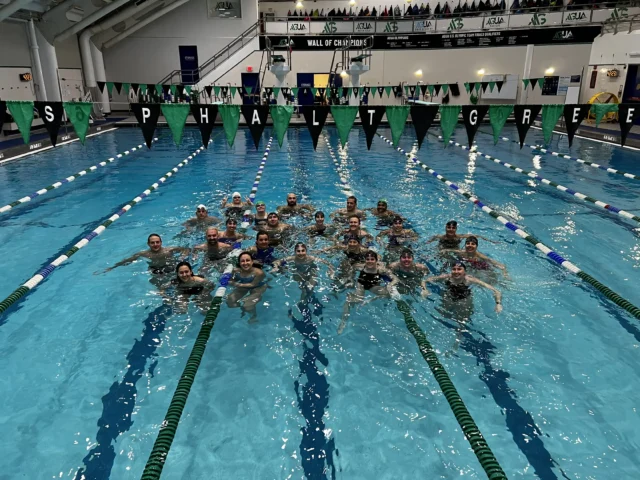 A group of swimmers posing together in an indoor swimming pool, with green and black pennant flags overhead and "ASPHALT GREEN" spelled out on the flags.