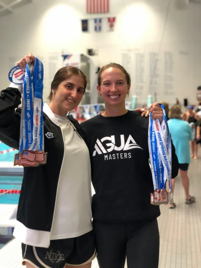 Two swimmers stand poolside, each holding multiple medals, smiling at the camera. A swimming pool and several people are visible in the background.