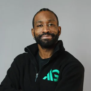A man with a beard and mustache wearing a black zip-up jacket with a green logo stands against a plain gray background, arms crossed and smiling slightly.