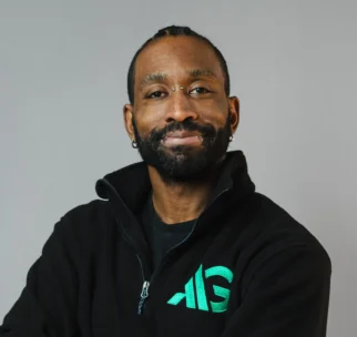 A man with a beard and mustache wearing a black zip-up jacket with a green logo stands against a plain gray background, arms crossed and smiling slightly.