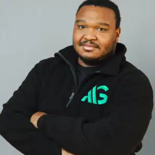 A man stands with arms crossed, wearing a black zip-up fleece with a green and teal "AIG" logo, in front of a plain gray background.