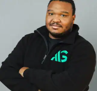 A man stands with arms crossed, wearing a black zip-up fleece with a green and teal "AIG" logo, in front of a plain gray background.