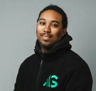 A man wearing a black zip-up jacket with a green AG logo stands against a plain gray background, facing the camera and smiling slightly.