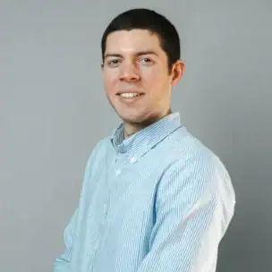 Man with short dark hair, wearing a light blue and white striped button-up shirt, smiles while standing against a plain light gray background.