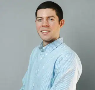 Man with short dark hair, wearing a light blue and white striped button-up shirt, smiles while standing against a plain light gray background.