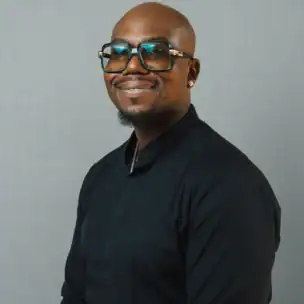 A man with glasses smiles at the camera. He is wearing a black long-sleeve shirt and standing against a plain, light gray background.