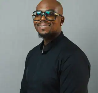 A man with glasses smiles at the camera. He is wearing a black long-sleeve shirt and standing against a plain, light gray background.