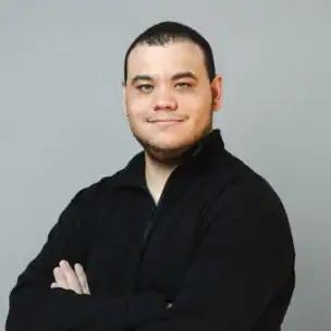 A person with short dark hair and a trimmed beard stands with arms crossed, wearing a black long-sleeve shirt, against a plain light gray background.