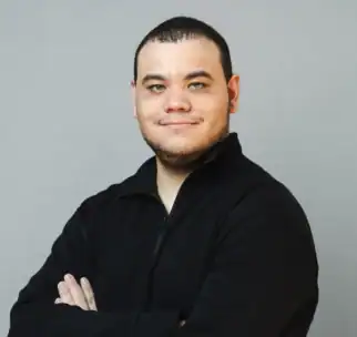 A person with short dark hair and a trimmed beard stands with arms crossed, wearing a black long-sleeve shirt, against a plain light gray background.
