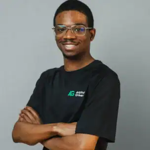 A person wearing glasses and a black "Alpha Green" t-shirt stands smiling with arms crossed against a plain gray background.