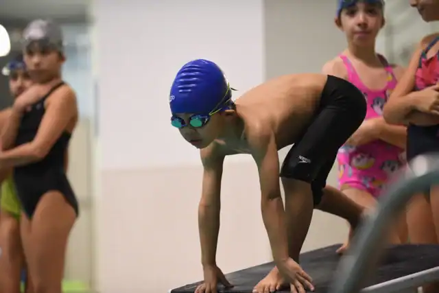 A young swimmer in a blue cap and goggles crouches on a starting block, ready to dive into the pool. Other swimmers are visible in the background.
