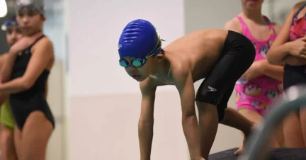 A young swimmer in a blue cap and goggles crouches on a starting block, ready to dive into the pool. Other swimmers are visible in the background.