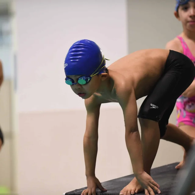 A young swimmer in a blue cap and goggles crouches on a starting block, ready to dive into the pool. Other swimmers are visible in the background.