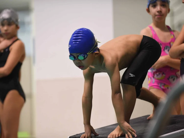 A young swimmer in a blue cap and goggles crouches on a starting block, ready to dive into the pool. Other swimmers are visible in the background.