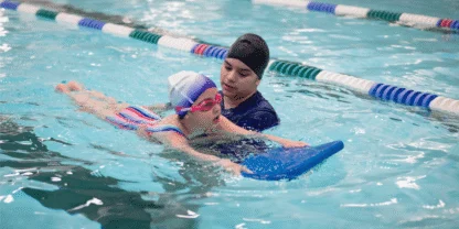 A swim instructor uses a blue kickboard to support a child in the water.