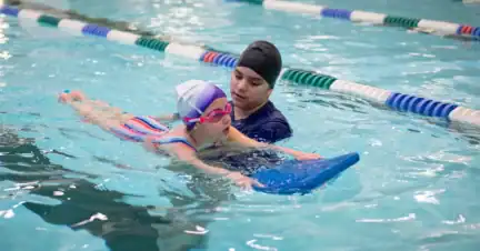 A swim instructor uses a blue kickboard to support a child in the water.