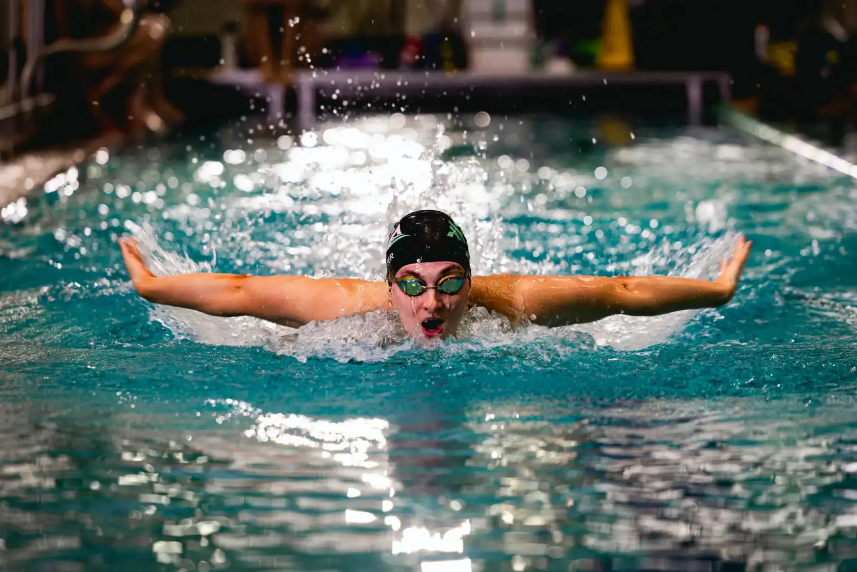 A swimmer wearing goggles and a swim cap performs the butterfly stroke in an indoor pool, with water splashing around their arms.