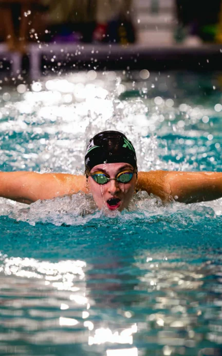 Swimmer wearing goggles and a swim cap performs the butterfly stroke in an indoor pool, water splashing around their arms.