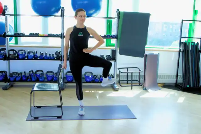 A person in athletic wear performs a standing exercise with one foot on a chair in a gym, surrounded by kettlebells, mats, and fitness equipment—an ideal setting for effective workout recovery.