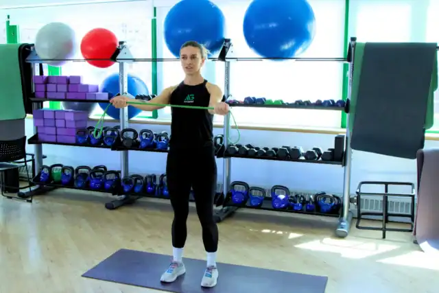 A person stands on a mat in a gym, holding a resistance band with arms extended forward, focusing on workout recovery. Gym equipment, weights, and exercise balls are visible in the background.