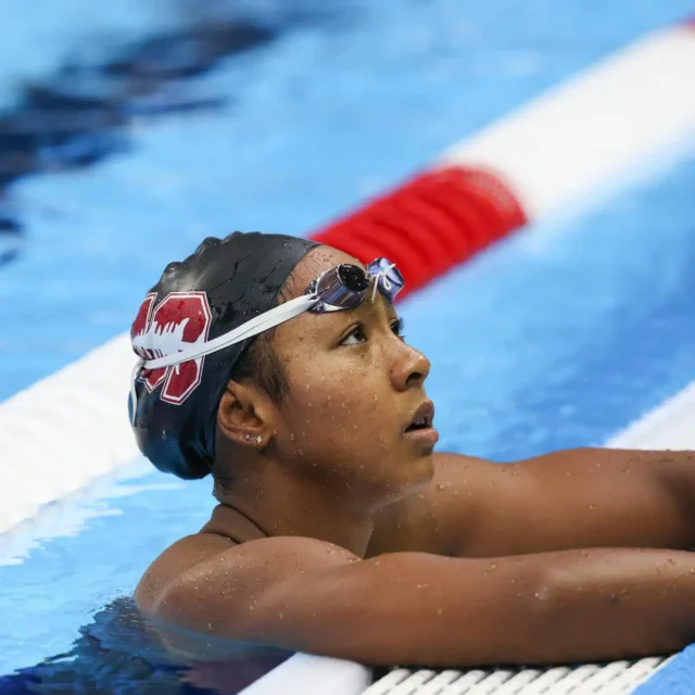 A swimmer wearing a black swim cap and goggles rests at the edge of an indoor pool, holding onto the poolside near a red and white lane divider.