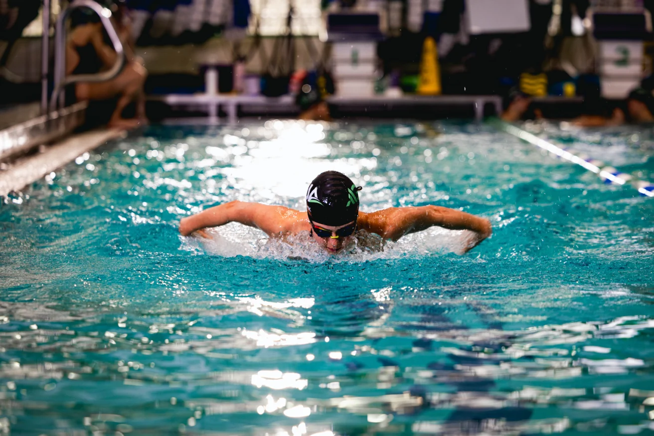 A swimmer wearing goggles and a cap performs the butterfly stroke in an indoor pool, creating splashes as they move forward.