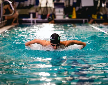 A swimmer wearing goggles and a cap performs the butterfly stroke in an indoor pool, creating splashes as they move forward.