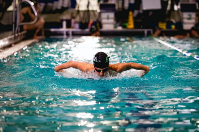 A swimmer wearing goggles and a cap performs the butterfly stroke in an indoor pool, creating splashes as they move forward.